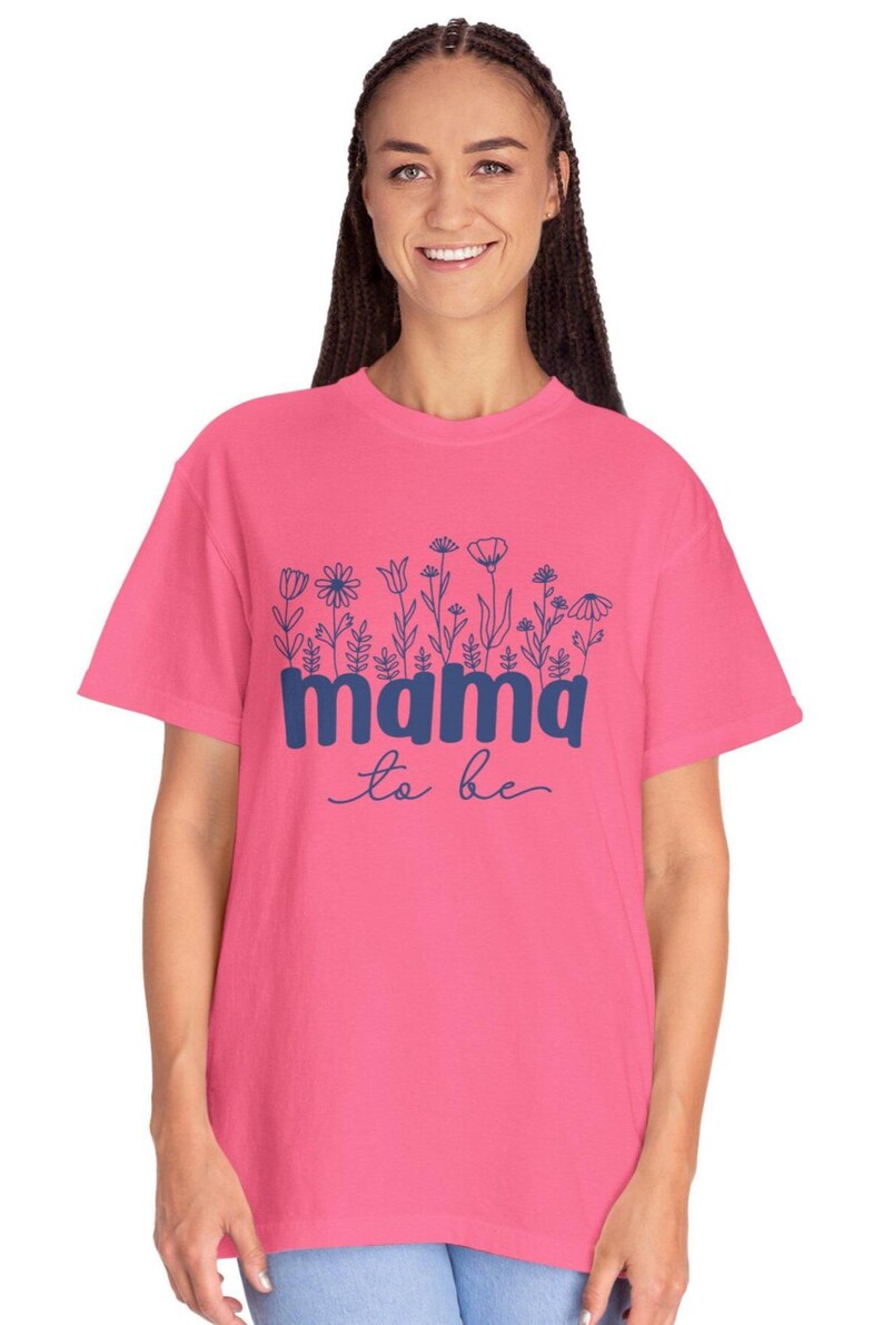 May include: A pink t-shirt with the words "mama to be" in blue script. The word "mama" is in a bold, blocky font, and the shirt features a floral design above the text. The shirt is a short-sleeved, crew neck style.