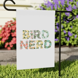 Puede incluir: Una bandera de jardín blanca con las palabras "BIRD NERD" en letras florales coloridas. La bandera se muestra en un soporte de metal negro en un jardín, con flores en el fondo.
