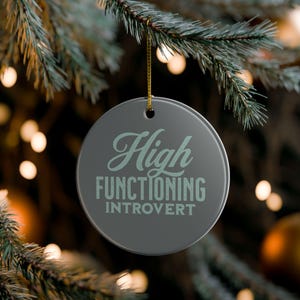 May include: A round, gray Christmas ornament with the words "High Functioning Introvert" in a light teal script. The ornament hangs from a gold string against a blurred background of Christmas tree branches and lights.