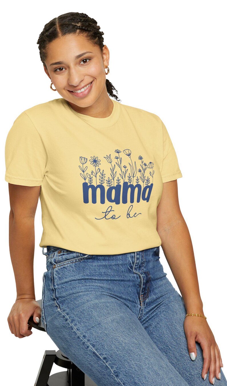 May include: A light yellow t-shirt with the text "mama to be" in blue, adorned with floral illustrations. The shirt is paired with blue jeans.