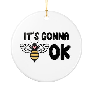 May include: White round ornament with gold string. The ornament features a cartoon bee with black wings and a yellow and black striped body. The text "IT'S GONNA OK" is printed in black above and to the right of the bee.