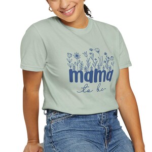 May include: Sage green t-shirt with a floral design and the words "mama to be" in navy blue. The shirt is a crew neck and is paired with blue jeans. The model is smiling.