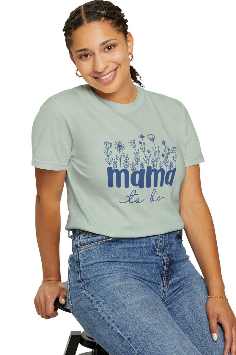 May include: Sage green t-shirt with a floral design and the words "mama to be" in navy blue. The shirt is a crew neck and is paired with blue jeans. The model is smiling.