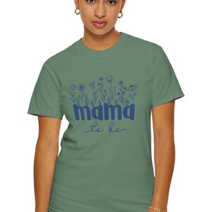 May include: Sage green t-shirt with a blue floral design and the words "mama to be". The shirt is made of a soft material and has short sleeves. The design includes various flower illustrations.