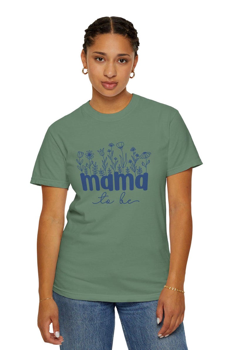 May include: Sage green t-shirt with a blue floral design and the words "mama to be". The shirt is made of a soft material and has short sleeves. The design includes various flower illustrations.