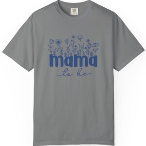 May include: Gray t-shirt with the words "mama to be" in blue, with a floral design above the word "mama". The shirt is a crew neck and has short sleeves. The text is in a script font.