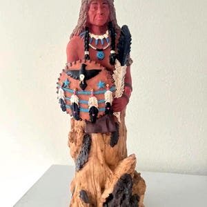 May include: A Native American figurine with a wolf headdress, holding a shield and feathers. The figure is painted in shades of red, brown, black, and blue. The shield features a bird and star design. The figurine is mounted on a wooden base.