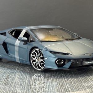 May include: A matte blue Lamborghini Temerario scale model car featuring a white racing stripe across the roof and side, intricate silver multi-spoke wheels, a low-profile aerodynamic body, and the word Temerario printed on the front bumper.