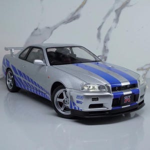 May include: Silver model car with blue racing stripes and accents. The car has a spoiler and the text "GT R" on the front. The car has a detailed design and is on a light grey surface.