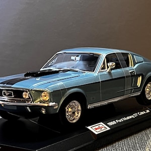 May include: A diecast model of a 1968 Ford Mustang GT Cobra Jet in a light blue color with black racing stripes. The model car sits on a black display stand. The model has chrome wheels and trim.