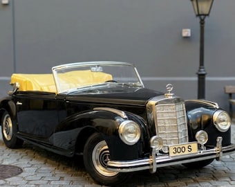 May include: A black vintage Mercedes-Benz 300 S convertible with a yellow top. The classic car has chrome accents, round headlights, and a front license plate that reads "300 S". The car is parked on a cobblestone street.