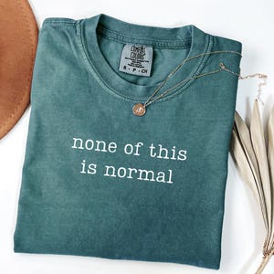 May include: A teal Comfort Colors t-shirt with the phrase "none of this is normal" in white lettering. A gold necklace with a small pendant rests on the shirt. The shirt is folded neatly.