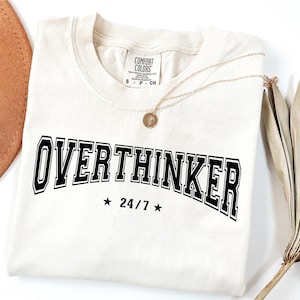 以下が含まれることがあります： 前面に「OVERTHINKER」の文字が黒色の太字で描かれたクリーム色のTシャツ。その下に「24/7」の文字が2つの星の間に配置されています。ペンダント付きのゴールドネックレスがシャツの上にかけられています。