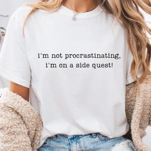 May include: White t-shirt with the text "i'm not procrastinating, i'm on a side quest!" in black. The shirt is made of a soft material and is being worn by someone. The shirt is paired with blue jeans.