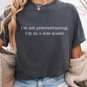 May include: Dark grey t-shirt with the text "I'm not procrastinating, I'm on a side quest!" in white. The shirt is a crew neck style and appears to be made of a soft, comfortable material. The model is wearing a necklace and a beige jacket.