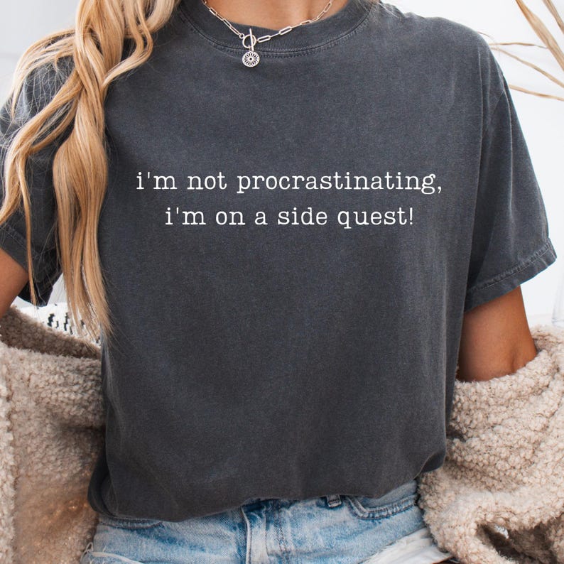 May include: Dark grey t-shirt with the text "I'm not procrastinating, I'm on a side quest!" in white. The shirt is a crew neck style and appears to be made of a soft, comfortable material. The model is wearing a necklace and a beige jacket.