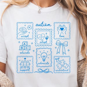 May include: White t-shirt with a blue graphic design featuring the word "autism" and related words like "accept," "adapt," "love," "educate," "advocate," "support," and "understand." The design includes puzzle pieces, a brain, and a heart.