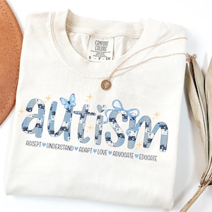 以下が含まれることがあります： クリーム色のTシャツ。青色のパッチワーク文字で「Autism」と書かれています。シャツには「Accept, Understand, Adapt, Love, Advocate, Educate」の文字が「Autism」の下に書かれています。ペンダント付きのゴールドネックレスがシャツの上にあります。