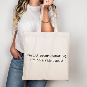 May include: Cream-colored tote bag with black text that reads, "I'm not procrastinating, I'm on a side quest!" The bag has two handles and is held up by a person. The person is wearing a white t-shirt and blue jeans.