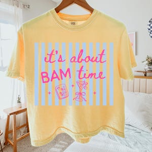 Puede incluir: Camiseta amarilla con la frase "it's about BAM time" en letras rosas. El dise&ntilde;o incluye un fondo a rayas azul claro, una copa de c&oacute;ctel y un peque&ntilde;o gr&aacute;fico rectangular. La camiseta est&aacute; colgada en una percha de madera.