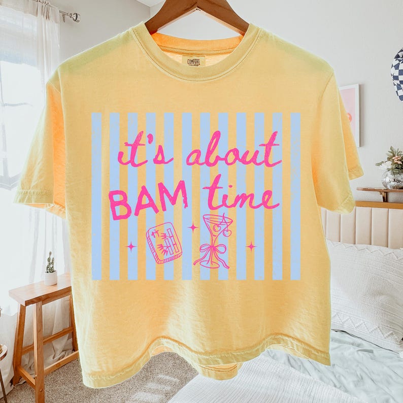 Puede incluir: Camiseta amarilla con la frase "it's about BAM time" en letras rosas. El dise&ntilde;o incluye un fondo a rayas azul claro, una copa de c&oacute;ctel y un peque&ntilde;o gr&aacute;fico rectangular. La camiseta est&aacute; colgada en una percha de madera.