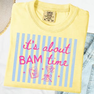 Puede incluir: Camiseta amarilla clara con el texto rosa "It's about BAM time" sobre rayas azules. El dise&ntilde;o incluye ilustraciones de un c&oacute;ctel y una pieza de juego.