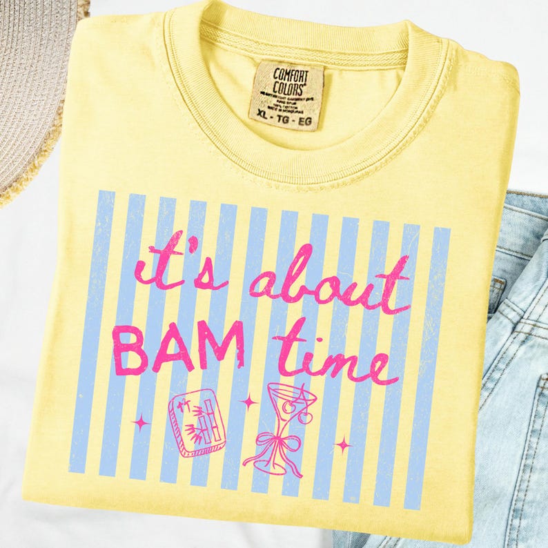 Puede incluir: Camiseta amarilla clara con el texto rosa "It's about BAM time" sobre rayas azules. El dise&ntilde;o incluye ilustraciones de un c&oacute;ctel y una pieza de juego.