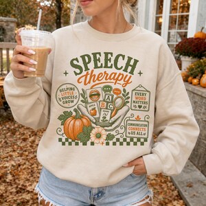 May include: A beige crewneck sweatshirt featuring a graphic print with the text "SPEECH therapy" in green and orange. The design includes illustrations of a pumpkin, alphabet blocks, a magnifying glass, and speech bubbles with text.