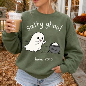 May include: An olive green crewneck sweatshirt featuring a white graphic of a ghost and a black pot labelled SALT. The text reads "salty ghoul" above the ghost and "i have POTS" below the pot, printed in a simple white sans-serif font.