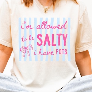 May include: Cream-colored t-shirt with the text "I'm allowed to be SALTY I have POTS" in pink lettering. The design includes a pink bow and light blue vertical stripes.