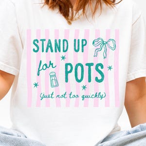 May include: A white t-shirt featuring a graphic with vertical light pink stripes and teal text that reads STAND UP for POTS (just not too quickly). The design includes a teal bow, a salt shaker icon, and small teal star accents.