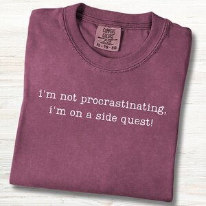 May include: A maroon t-shirt with the text "i'm not procrastinating, i'm on a side quest!" in white. The shirt has a crew neck and a tag that reads "COMFORT COLORS XL-TG-EG". The shirt is folded neatly.