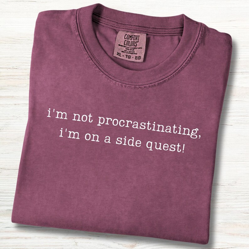 May include: A maroon t-shirt with the text "i'm not procrastinating, i'm on a side quest!" in white. The shirt has a crew neck and a tag that reads "COMFORT COLORS XL-TG-EG". The shirt is folded neatly.