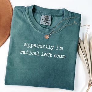 Op de afbeelding: Een teal Comfort Colors t-shirt met de tekst "apparently i'm radical left scum" in het wit. Een gouden ketting met een hanger en een bruine hoed zijn ook zichtbaar.