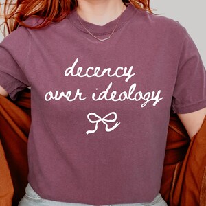 Op de afbeelding: Een pruimkleurig t-shirt met korte mouwen en de witte tekst "decency over ideology" in een handgeschreven lettertype, geplaatst boven een eenvoudige witte lijntekening van een strik in het midden van de borst van het kledingstuk.