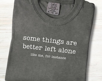 May include: A folded, dark gray t-shirt with the text "some things are better left alone like me, for instance" in white. The shirt has a Comfort Colors tag in the upper right corner.