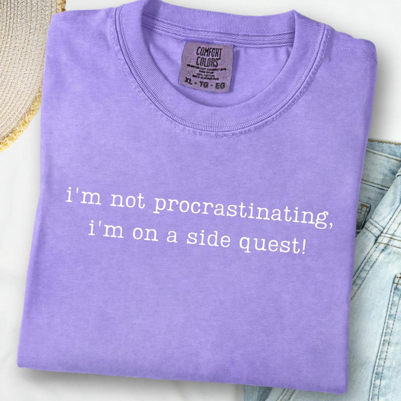 May include: A lavender Comfort Colors t-shirt with the text "i'm not procrastinating, i'm on a side quest!" in white. The shirt is folded and has a tag in the upper right corner. The shirt is on a white surface.