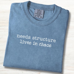 May include: A folded, light blue t-shirt with the text "needs structure lives in chaos" in white. The shirt has a crew neck and a tag that reads "Comfort Colors". The shirt is on a light wood surface.
