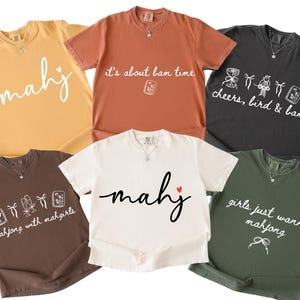 Könnte beinhalten: Sechs kurzärmelige T-Shirts in verschiedenen Farben, darunter Gelb, Orange, Schwarz, Braun, Weiß und Grün. Jedes Shirt hat ein anderes weißes Grafikdesign und Text zum Thema Mahjong.