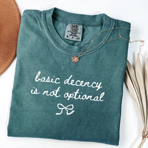 Op de afbeelding: Een teal Comfort Colors t-shirt met de tekst "basic decency is not optional" in witte letters. Een gouden ketting met een kleine hanger ligt op het shirt. Het shirt is opgevouwen en een bruine hoed is gedeeltelijk zichtbaar op de achtergrond.
