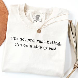 May include: Cream-colored t-shirt with the text "I'm not procrastinating, I'm on a side quest!" in black. A gold necklace with a pendant rests on the shirt. The shirt has a Comfort Colors tag.