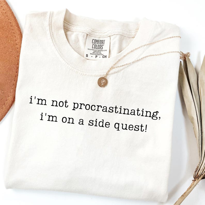 May include: Cream-colored t-shirt with the text "I'm not procrastinating, I'm on a side quest!" in black. A gold necklace with a pendant rests on the shirt. The shirt has a Comfort Colors tag.