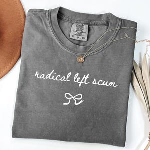 Op de afbeelding: Een grijs Comfort Colors T-shirt met de woorden "radical left scum" in witte letters en een strik-afbeelding. Een gouden ketting met een hanger ligt over het shirt.