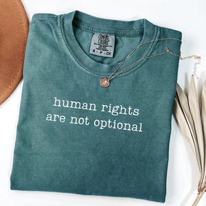 Op de afbeelding: Een teal Comfort Colors t-shirt met de tekst "human rights are not optional" in het wit. Een gouden ketting met een hanger is over het shirt gedrapeerd. Een bruine hoed en een gedroogde plant staan op de achtergrond.