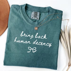 以下が含まれることがあります： 「bring back human decency」の白い文字が書かれた、ティール色のComfort Colors Tシャツ。ペンダント付きのゴールドネックレスがシャツの上にあります。茶色の帽子と乾燥した植物が背景にあります。