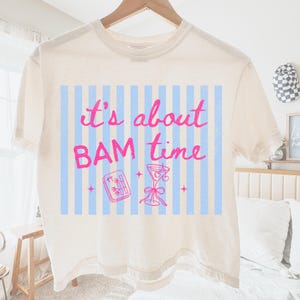 Puede incluir: Camiseta color crema con la frase "it's about BAM time" en rosa. El dise&ntilde;o incluye un fondo a rayas azules y blancas, un c&oacute;ctel y una ilustraci&oacute;n de rollo de sushi de bamb&uacute;.