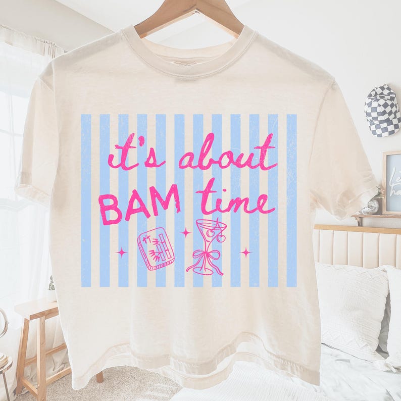 Puede incluir: Camiseta color crema con la frase "it's about BAM time" en rosa. El dise&ntilde;o incluye un fondo a rayas azules y blancas, un c&oacute;ctel y una ilustraci&oacute;n de rollo de sushi de bamb&uacute;.