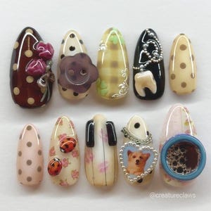 Puede incluir: Una colección de diez uñas artificiales con varios diseños. Las uñas presentan lunares, flores, una cara sonriente, un diente, mariquitas, un piano, un perro en un corazón y una taza de café. Las uñas son de diferentes colores, incluyendo marrón, beige, negro y rosa.