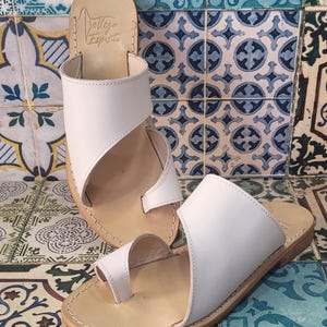 May include: Two pairs of white leather sandals with a toe loop and a strap across the foot. The sandals have a brown leather sole and are on a tiled background.