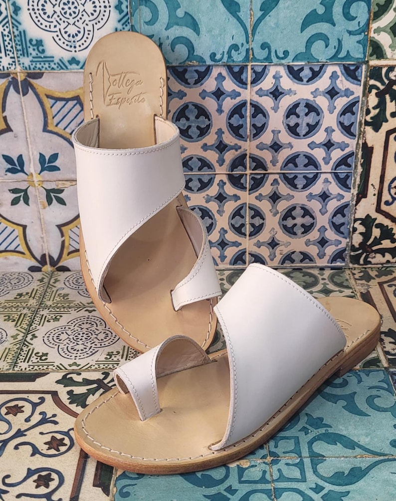 May include: Two pairs of white leather sandals with a toe loop and a strap across the foot. The sandals have a brown leather sole and are on a tiled background.